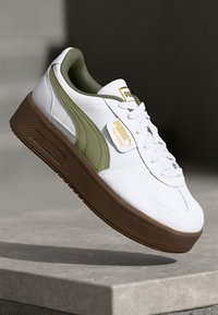 Zapatilla blanca con detalles en verde oliva y suela de goma marrón texturada. Presenta un diseño clásico de cordones y una etiqueta de logo prominente.
