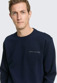 Granatowa bluza wykonana z miękkiej bawełny, z okrągłym dekoltem i małym logo na piersi. Prosty, klasyczny krój.