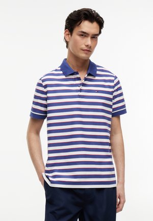 Bărbat tânăr îmbrăcat cu un tricou polo cu dungi albastre, albe și roșii, cu guler albastru și pantaloni bleumarin, stând în fața unui fundal alb simplu.