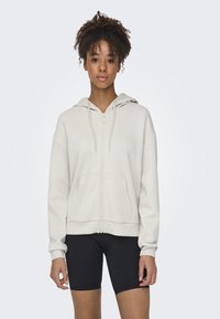 Light beige zip-up hoodie made of soft fabric, featuring a front pocket, ribbed cuffs, and a hood with drawstrings. Black shorts worn underneath.