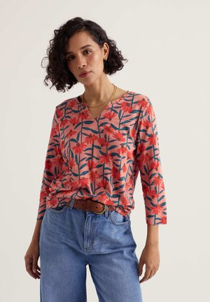 Žena v koralovo ružovom kvitnutom tričku s dlhými rukávmi a modrých džínsach s hnedým opaskom stojaca na svetlom jednofarebnom pozadí.