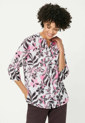Frau mit lockigem Haar, lächelnd, trägt eine weiße Bluse mit rosa und schwarzen Blumenmustern und dunkle Hose, Hand berührt Hals und Hüfte.
