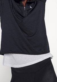 Svart zippered träningsoverdel med draperad front, lager på lager över en ljusgrå mesh-tröja, parat med svarta leggings, slät textur genomgående.