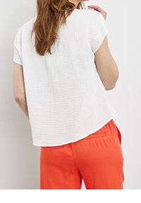 Femme aux longs cheveux bruns portant un haut blanc texturé à manches courtes et un pantalon orange vif, se tenant devant un fond blanc.