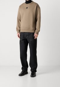 Persona che indossa una felpa beige a maniche lunghe con uno strato di collo nero, pantaloni di jeans neri con grandi tasche frontali e scarpe di pelle nere.