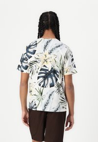 Kortermet tropisk skjorte med hvit bakgrunn, med blå og gule blomstermønstre, kombinert med mørkebrune shorts.