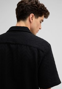 Strukturierte schwarze Kurzarmbluse mit einem Fischgrätmuster und Kragen, von hinten gezeigt mit sichtbaren Nähten und einem lockeren Schnitt.