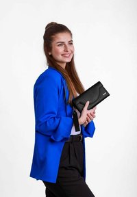 Černá kožená peněženka s zakřiveným klopou, která má tři dekorativní hřebíky. Držena proti osobě v modrém saku a černých kalhotách.