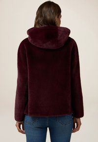 Giacca cappuccio bordeaux in tessuto morbido e lussuoso. Presenta una vestibilità rilassata ed è abbinata a jeans blu aderenti.