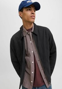 Giacca nera realizzata in materiale liscio con zip frontale, indossata sopra una camicia a quadri rossa e bianca, strato sopra una maglietta bordeaux.