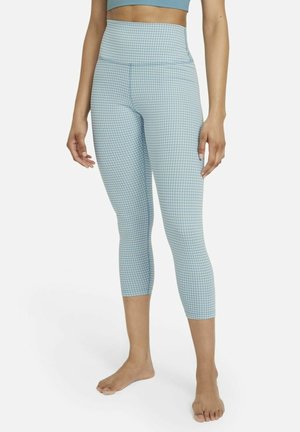 Mujer con leggings cortos de cintura alta de cuadros azules y camiseta corta azul claro, de pie descalza sobre un fondo blanco.