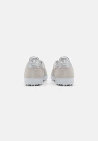 Sneakers din suede beige cu accente argintii, având un tab de călcâi texturat și tălpi din cauciuc alb. Branding vizibil în zona călcâiului.