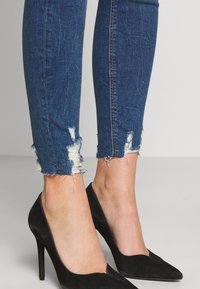 Jeans ajustados de denim azul oscuro con dobladillo desgastado y bordes deshilachados, combinados con zapatos de tacón alto negros. Textura suave, diseño ceñido.