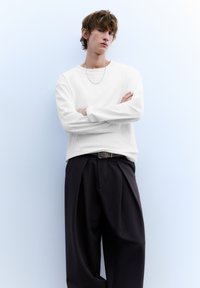 Weißes Langarmshirt mit lockerem Schnitt, kombiniert mit schwarzen, plissierten Hosen. Accessoires umfassen eine dünne silberne Kettenhalskette und einen Gürtel.