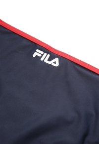 Marineblauwe stof met een rode bies bevat het witte "FILA" logo. Het materiaal lijkt glad en lichtgewicht, geschikt voor sportkleding.