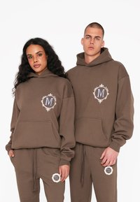 Brune hættetrøjer med frontlomme og matchende joggingbukser. Hver af dem har et broderet logo inden for en dekorativ ramme. Blød tekstur.