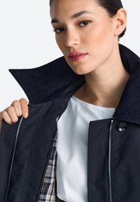 Juoda striukė su tekstūruotu tamsiu apykakliu, užtrauktuku užsegama ir languotu pamušalu; po apačia matyti dalis balto marškinio.