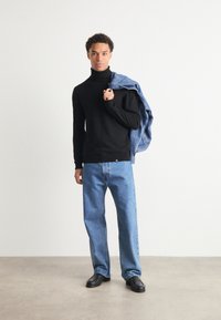 Μαύρος πουλόβερ με μανίκια τσέπης, χαλαρά μπλε τζιν και μαύρες λοφάνοι. Το μοντέλο κρατάει ένα  τζην μπουφάν πάνω από τον ώμο. Απλός σχεδιασμός και ήσυχες αποχρώσεις.