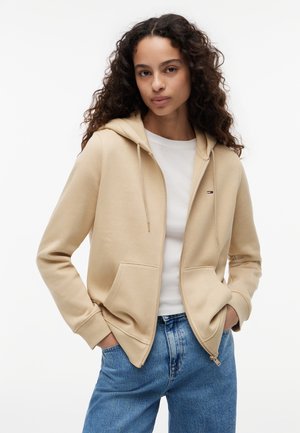 Młoda kobieta o kręconych włosach, ubrana w beżową bluzę z zamkiem na suwak na białej koszulce i niebieskich dżinsach, dłonie w kieszeniach bluzy, neutralne tło.