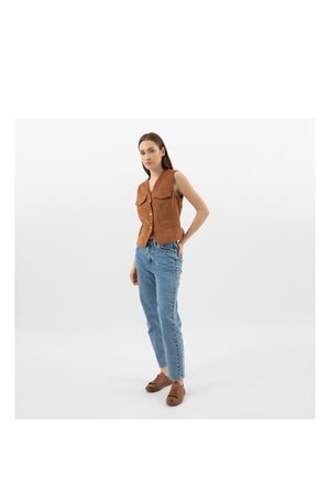 Mladá žena stojící s jednou rukou v kapse, oblečená v bezrukávové hnědé vestě na knoflíky, světlých modrých džínách a hnědých teniskách, na jednobarevném bílém pozadí.
