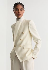 Žena s pletenými vlasmi, nosiaca dvojradové béžové sako so zlatými gombíkmi a zladené nohavice, pozerá sa do strany.