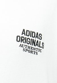 Λευκό ύφασμα με μαύρα κεντημένα γράμματα που γράφουν «ADIDAS ORIGINALS AUTHENTIC SPORTS» με κεφαλαία γράμματα.