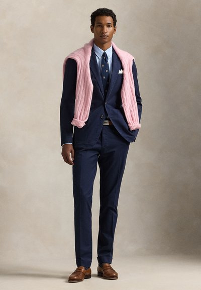 Mladý muž oblečený v námořnicky modrém obleku a pruhované košili s vzorovanou kravatou, růžový svetr přehozený přes ramena a hnědé kožené boty.