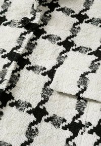 Tejido de tweed en blanco y negro texturizado con un patrón geométrico y un acabado suave, acentuado por un botón negro y un bolsillo con solapa.