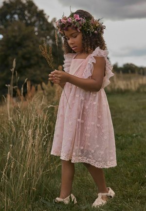 Jeune fille en robe rose en dentelle et couronne de fleurs tenant des herbes sauvages, debout dans un champ d'herbe sous un ciel nuageux.