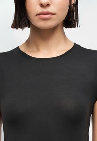 Γυναίκα με κοντά σκούρα μαλλιά που φορά απλό μαύρο t-shirt με στρογγυλή λαιμόκοψη, εμφανιζόμενη από τους ώμους έως τα χείλη σε λευκό φόντο.