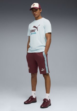 Hombre con camiseta Puma Portugal azul claro, pantalones cortos granate con franjas laterales estampadas, gorra a juego, calcetines blancos y zapatillas granates.