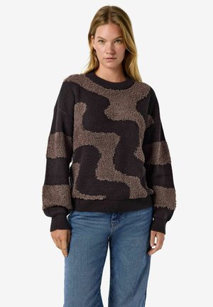 Mulher vestindo um casaco preto e castanho com padrão texturizado, com jeans azuis, em pé contra um fundo branco simples.