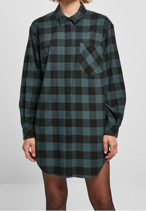 Femme portant une robe-chemise ample à manches longues aux carreaux noirs et vert foncé, avec une poche avant et un col boutonné.