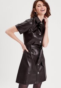 Robe chemise en simili cuir noir avec des manches courtes, fermeture à boutons sur le devant, taille ceinturée et jupe évasée. Motif texturé sur les collants.