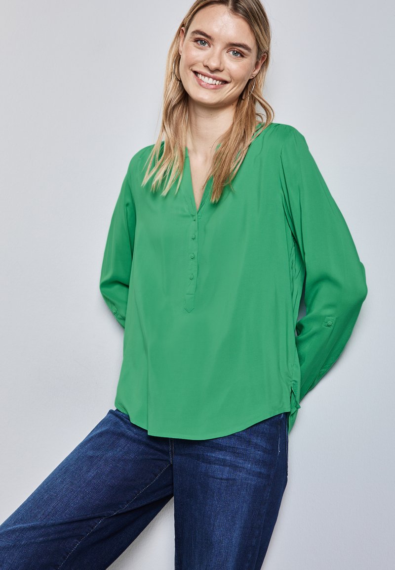 Blouse verte à manches longues avec un décolleté en V, présentant des boutons à l'avant, fabriquée en tissu lisse, associée à un jean bleu.