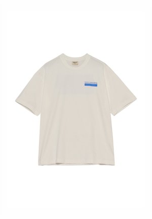 Ung mand står fremadrettet iført en løs hvid T-shirt med blå tekst og beige cargobukser med sneakers på hvid baggrund.