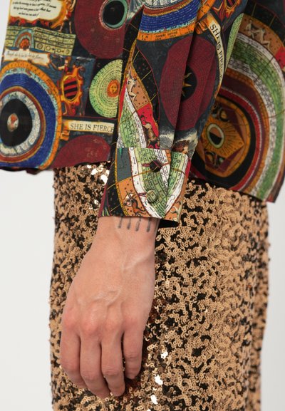 Multikleurige shirt met patronen en abstracte ontwerpen en tekst, gecombineerd met een glimmende gouden paillettenrok. Bevat een nauwsluitende manchet en zichtbare handtattoo.