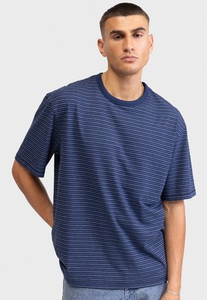 Tânăr care poartă un tricou bleumarin cu mâneci scurte și dungi subțiri și blugi de culoare albastru deschis, stând în picioare într-un cadru simplu, cu fundal deschis.