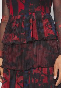 Gros plan sur un tissu plissé avec des volants superposés dans un motif floral abstrait rouge, noir et bleu sur un vêtement à manches longues.