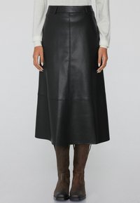Falda midi de cuero negra con una cinturilla ajustada, detalles en las costuras y una silueta ligeramente acampanada, combinada con botas marrones hasta la rodilla.