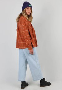 Chemise à carreaux rouille avec des boutons, associée à un jean large bleu clair et des bottines noires. Bonnet en tricot violet sur la tête.