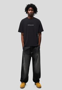 Tricou negru supradimensionat cu text alb „CHAOTICALLY CUTE”, asortat cu blugi negri cu picioare largi și bocanci de muncă galbeni. Design simplu, modern.