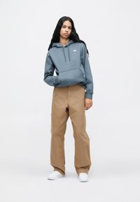 Νεαρή γυναίκα με μακριά μαλλιά, ντυμένη με μπλε φούτερ, καφέ παντελόνι φαρδύ και λευκά αθλητικά παπούτσια, που στέκεται μπροστά σε έναν απλό ανοιχτό φόντο.