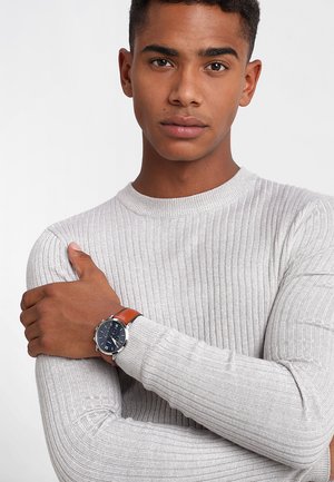 Montre à bracelet en cuir marron avec un boîtier rond en argent et un cadran bleu, arborant des index et des aiguilles en argent, portée au poignet d'un modèle.