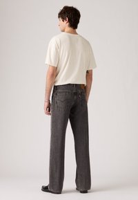 Man met kort donker haar, gekleed in een witte t-shirt, losse donkergrijze jeans en zwarte schoenen, staand met zijn rug naar de camera tegen een effen achtergrond.