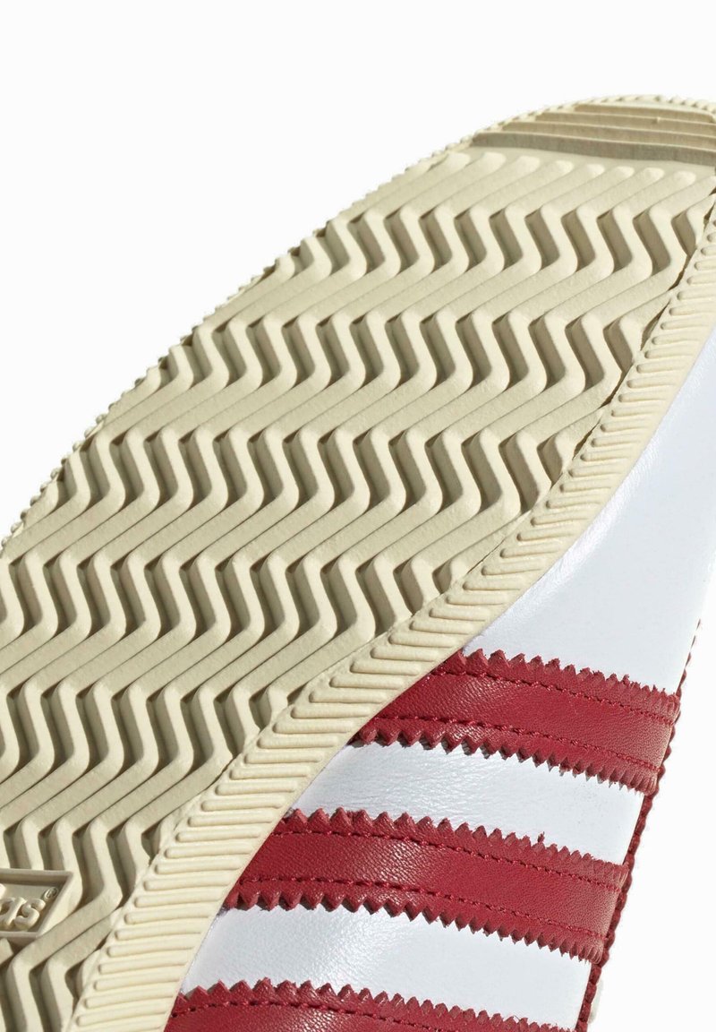 Weißer Sneaker-Sohle mit roten Streifen, strukturiertem Gummimuster und einer geschwungenen Form. Enthält eine Zickzack-Kantenverzierung und eine cremefarbene Außensohle.