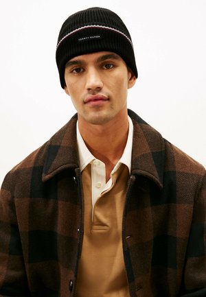 Beanie a coste nera con un accento a strisce orizzontali e toppa con logo, indossato con una giacca a quadri marrone e nera sopra una maglietta beige.