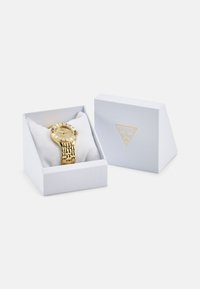 Goldene Uhr mit rundem Ziffernblatt, strukturiertem Armband, präsentiert in einer weißen Schachtel mit weichem Innenraum. Die Schachtel hat ein goldenes Logo auf der Vorderseite.