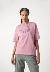 Roza bombažna majica s kratkimi rokavi in okroglim izrezom. Na sprednji strani je v temno sivi barvi natisnjeno "NEW BALANCE 1906". Sproščen kroj.