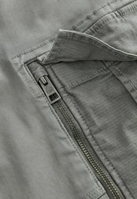 Tejido gris con una textura suave presenta un cierre de cremallera con una lengüeta negra. Las costuras a lo largo del borde añaden un detalle al bolsillo.